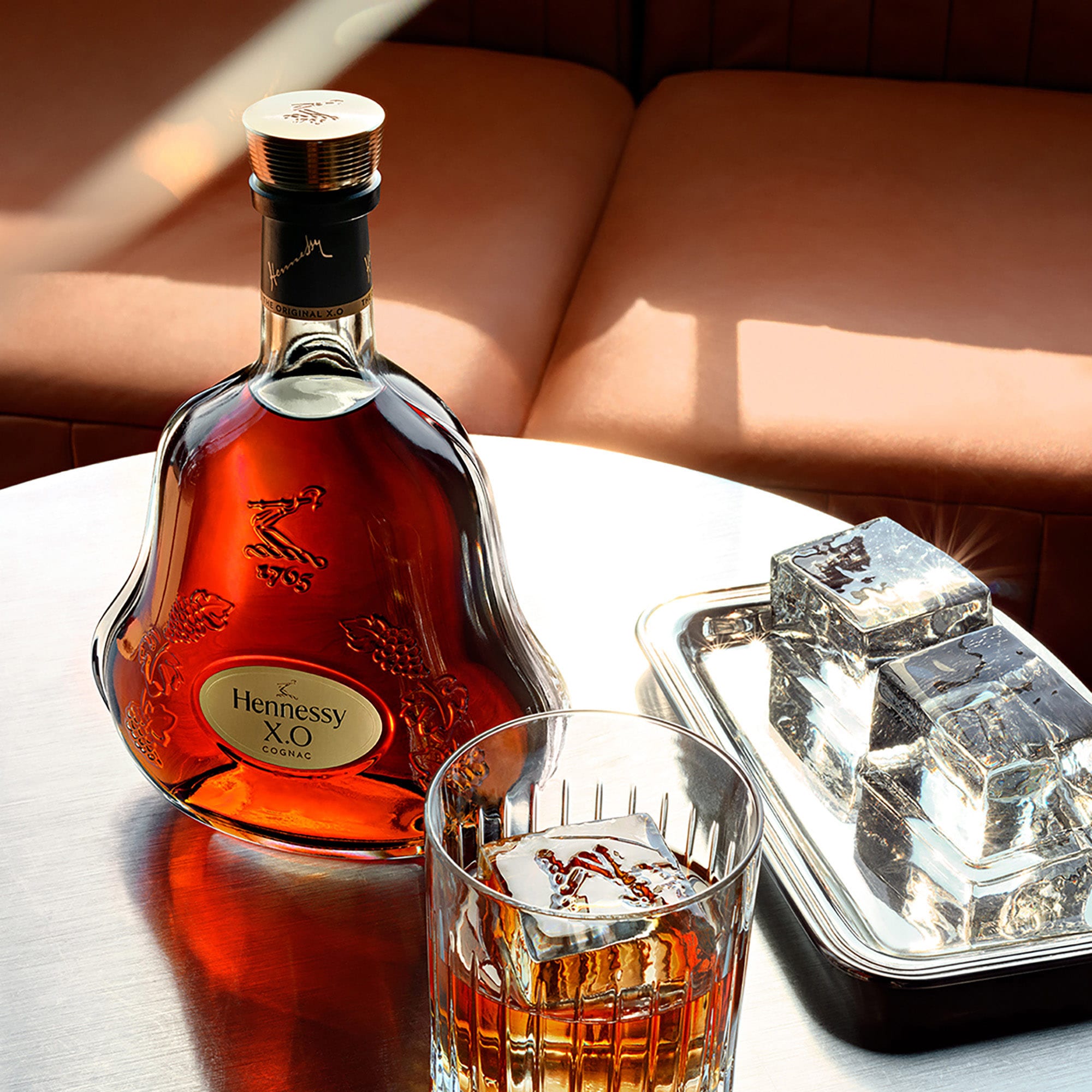 Decanter of Hennessy X.O next to a glass of cognac and large ice cubes, on a coffee table